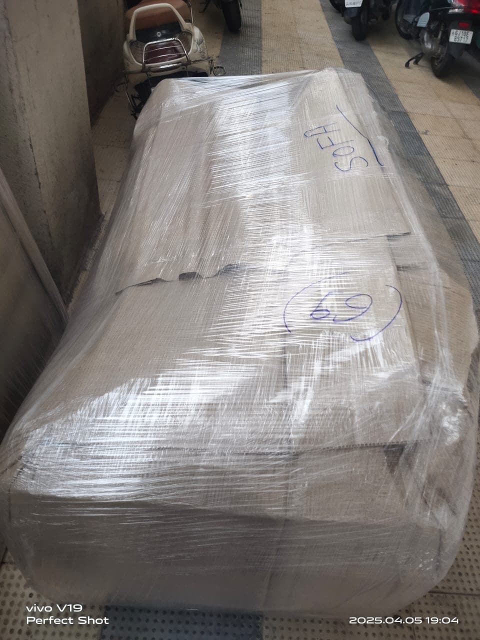 A large sofa wrapped in protective film beside parked scooters on a tiled walkway