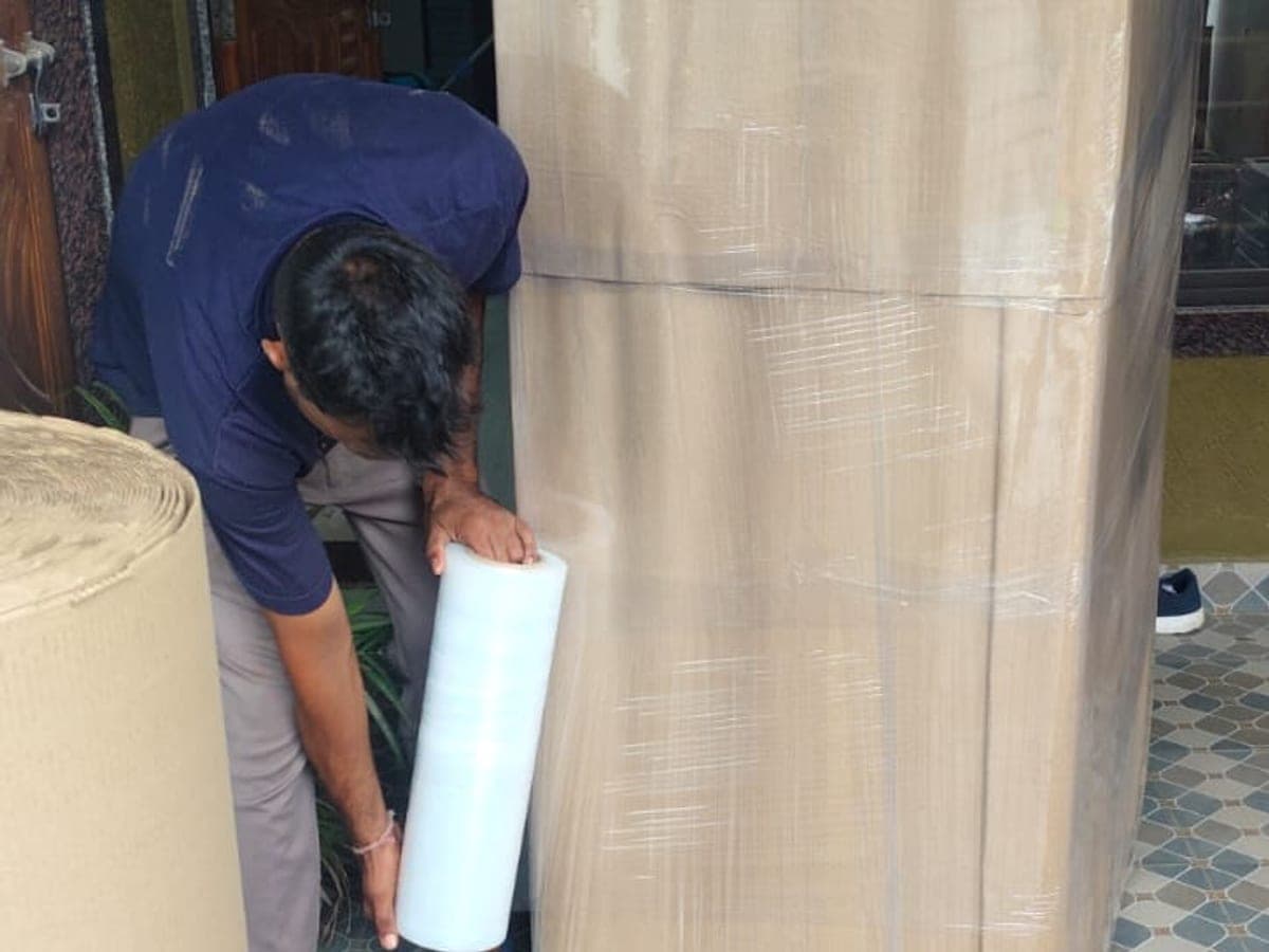 A mover wrapping a refrigerator in stretch film at a doorway, with corrugated rolls alongside