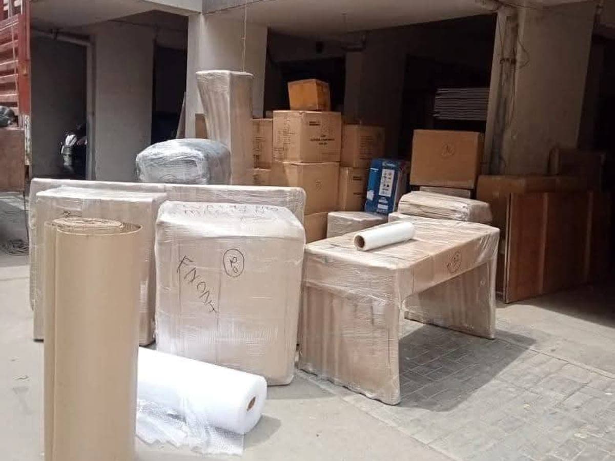 Packed cartons, wrapped tables and rolls of corrugated sheet staged outside a building before loading