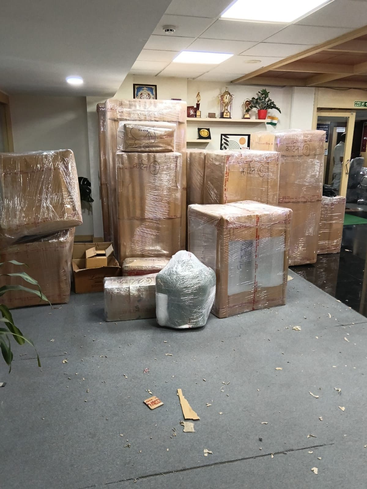 Stacked and numbered cartons wrapped in plastic inside an office lobby with trophies on a shelf behind