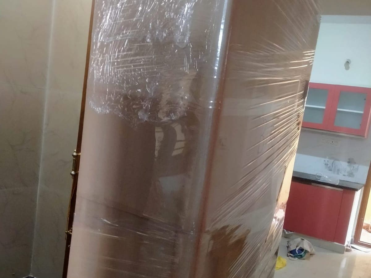 A tall wardrobe wrapped in stretch film inside a home, with a kitchen visible in the background